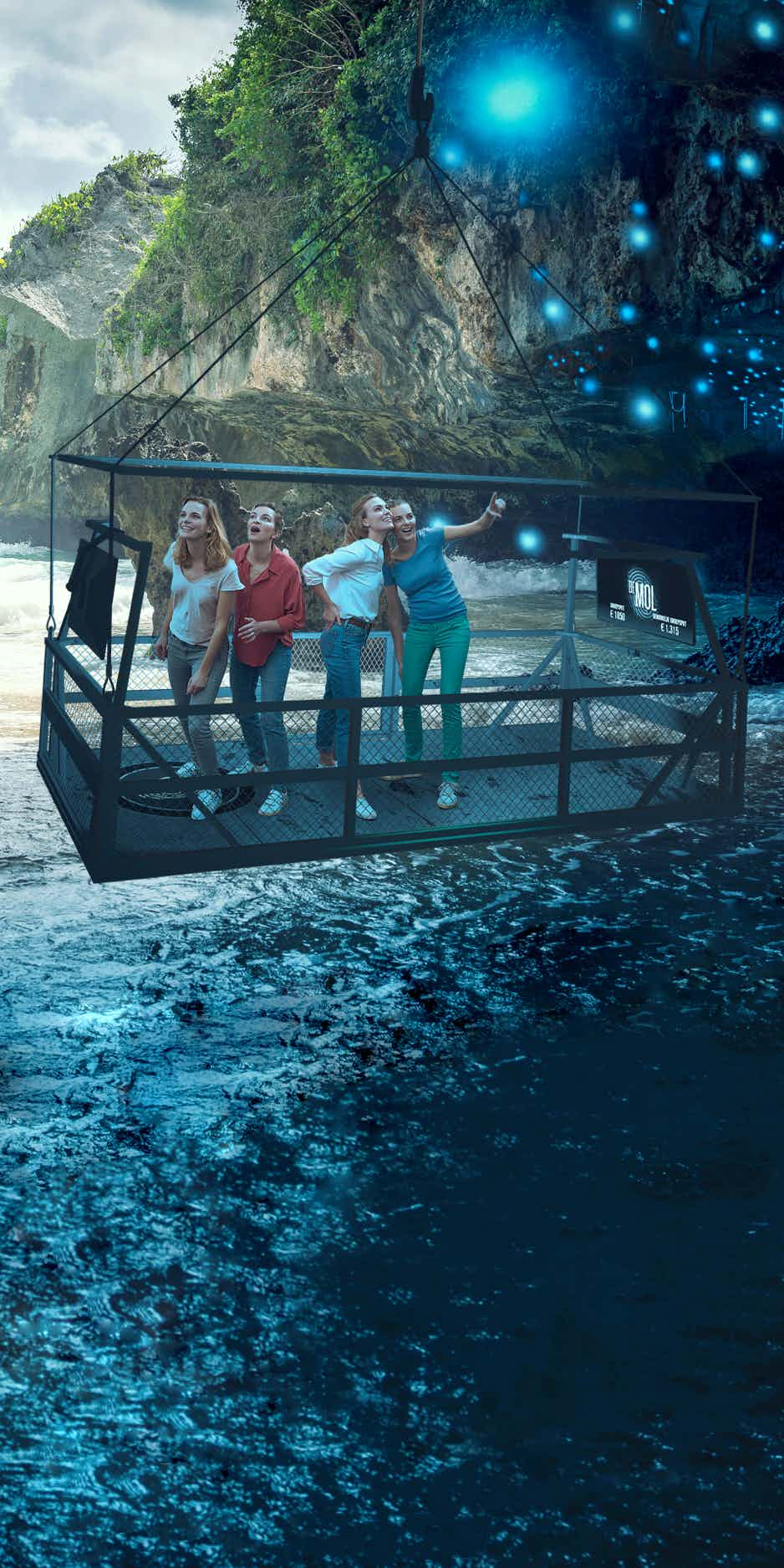 Girls standing on platform hovering over water in a cave, looking in wonder at the blue fireflies.